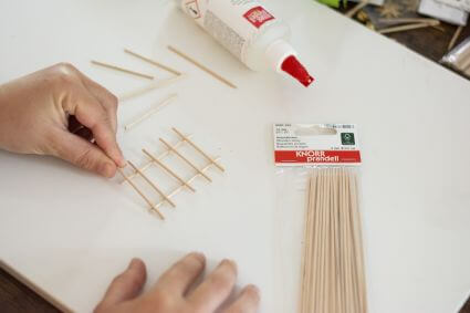 Blick auf einen weißen Schreibtisch, wo zwei Hände mit Hilfe von Bastelhölzern von Knorr Prandell eine kleine Leiter für eine selbstgemachte Wichteltür bauen.