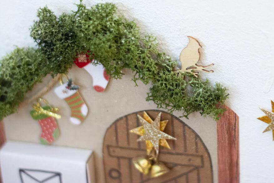 Nahaufnahme einer selbstgebastelten Wichteltür: Ein Holzornament in Form eines Vogels sitzt auf dem mit Moos bewachsenen Dach des Wichtelhauses.