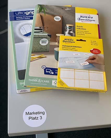 Auf einem Tisch liegen mehrere Verpackungen mit Etiketten der Marke Avery Zweckform. Der Tisch ist mit einem runden Etikett mit den Worten Marketing 1 beschriftet.