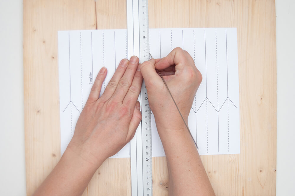 Pappvase: Jemand fährt auf einem Designpapier von Artoz die Linien einer Faltvorlage mit einer dünnen Stricknadel entlang eines Lineals nach.