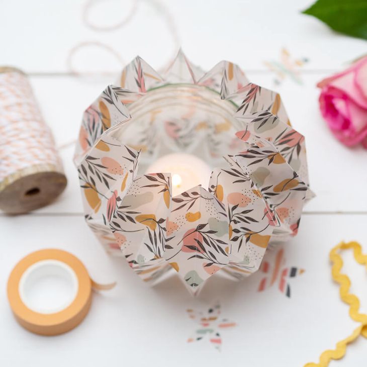 Ein gefaltetes Windlicht aus Transparentpapier der Marke Artoz steht auf einem weißen Untergrund. Drumherum sind verschiedene Bastelmaterialien verteilt.