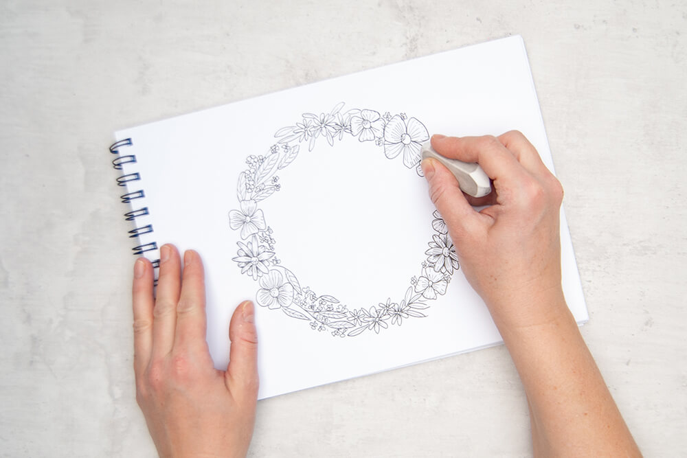 Jemand entfernt einen gezeichneten Bleistift-Kreis mit einem Radiergummi von Staedtler. Zurück bleibt ein Pigment Liner Blumenkranz auf einem Malblock. Foto: Johanna Rundel