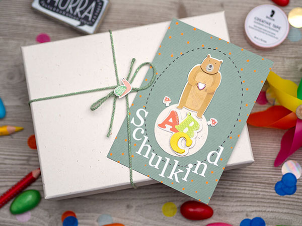 Auf einem hellgrauen Holzuntergrund steht eine weiße Geschenkschachtel aus Pappe. Darauf liegt eine wasserblaue Grußkarte, auf die ein brauner Bär, die Buchstaben ABC und das Wort Schulkind mit Aufklebern der Firma Rössler geklebt ist. Drumherum liegen buntes Konfetti, eine Garnspule, Washitape und verschiedene Stifte verteilt.