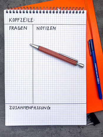 Auf einem dunklen Untergrund liegt ein orangefarbenes Notizbuch, darauf ein karierter Notizblock. Auf dem Block ist die Aufteilung für die Cornell Methode gezeichnet. Ein Kugelschreiber und ein Fasermaler der Marke Staedtler liegen auf und neben dem Block.