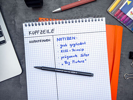 Auf einem dunklen Untergrund liegt ein orangefarbenes Notizbuch, darauf ein karierter Notizblock. Auf dem Block ist die Aufteilung für die Cornell Methode gezeichnet. Der Bereich für die Notizen ist mit blauem Stift farblich hervorgehoben. Auf dem Block liegt ein Fasermaler der Marke Staedtler. Drumherum ist weiteres Büromaterial verteilt.