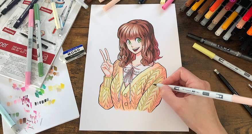 Jemand koloriert mit einem Marker ein Manga. Dieses zeigt eine junge Frau mit braunen Haaren und einem gelben Pulli, die lachend die rechte Hand mit dem Peace-Zeichen hochhält. Drumherum sind viele Zeichenmaterialien der Marke Tombow verteilt. Foto: Reyhan Yildirim