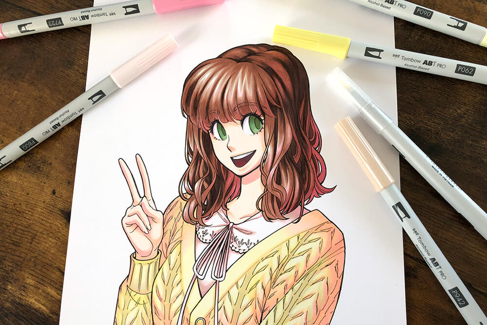 Auf einem braunen Holztisch liegt ein Blatt Papier. Darauf ist die Fineliner-Skizze einer jungen Frau im Manga-Stil zu sehen, die gerade mit Markern von Tombow koloriert wurde. Foto: Reyhan Yildirim