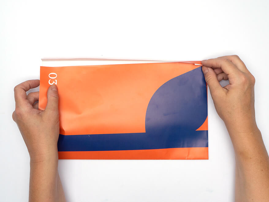 Tüte aus Papier falten ohne Vorlage: Jemand faltet ein rechteckiges, orangefarbenes Kalenderblatt von Athesia mittig zusammen. Der Untergrund ist weiß. Foto: Johanna Rundel
