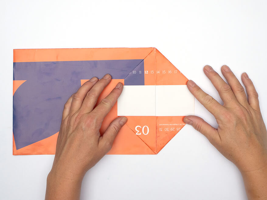 Tüte aus Papier falten ohne Vorlage: Jemand faltet ein Stück eines rechteckigen, orangefarbenen Kalenderblatts von Athesia, das vorgefaltet wurde, diagonal auseinander. Der Untergrund ist weiß. Foto: Johanna Rundel