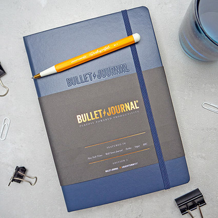 Auf einem hellgrauen Untergrund liegt ein dunkelblaues Bullet Journal von Leuchtturm1917. Darauf liegt ein orangefarbener Drehgriffel. Einige weiße Büroklammern und Foldback-Klammern liegen verstreut herum. Im Hintergrund steht ein blaues Glas mit Wasser. Foto: Johanna Rundel 