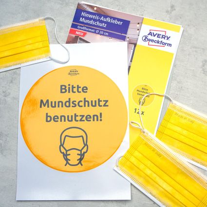 Auf einem grauen Untergrund in Betonoptik liegt eine Packung Hinweis-Aufkleber zum Thema Mundschutz von Avery-Zweckform. Darauf liegt ein Bogen mit dem gelben Hinweisaufkleber. Rundherum liegen ein paar gelbe Mund-Nasen-Bedeckungen. Foto: Johanna Rundel