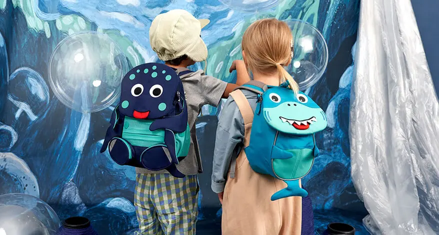 Zwei Kindergartenkinder stehen mit dem Gesicht vor einer bemalten Wand. Sie haben Kindergarten-Rucksäcke der Marke Affenzahn in Form eines Hais und Fischs auf dem Rücken.