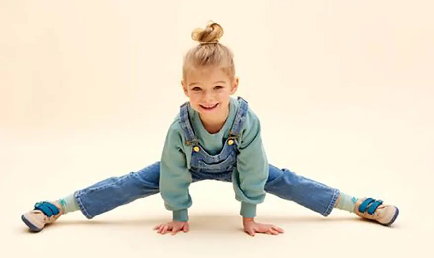 Ein blondes Mädchen mit Latzhose übt einen Spagat.