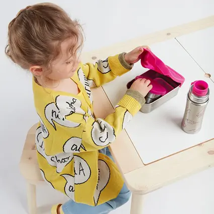 Ein Mädchen in einem gelben Pullover packt am Tisch seine Edelstahlbrotdose mit pinkem Deckel von Affenzahn aus. Daneben steht eine passende Trinkflasche.