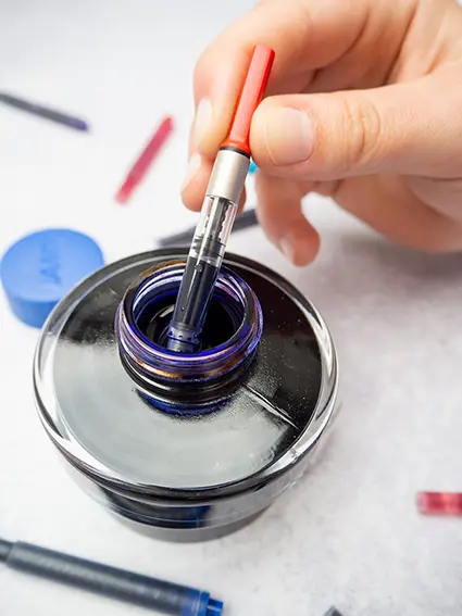 Eine Hand zieht Tinte aus einem Tintenfass in einen roten Konverter von Lamy auf. Drumherum liegen bunten Tintenpatronen verstreut.