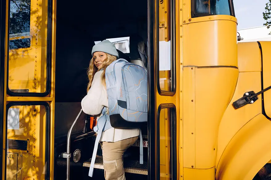 Ein Mädchen mit einem hellblauen Schulrucksack von satch steigt in einen gelben Schulbus ein.