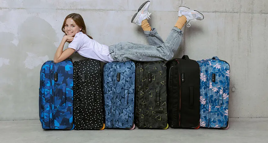 Ein jugendliches Mädchen liegt auf einer Reihe Rollkoffern in Blautönen von satch. Sie hat den Kopf in die Arme gestützt und lacht fröhlich in die Kamera. Der Hinter- und Untergrund ist aus Beton.