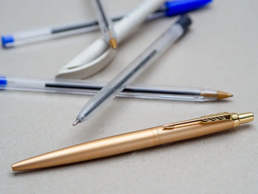 Auf einem grauen Untergrund liegen im Hintergrund einige billige Einweg-Kugelschreiber. Im Vordergrund ist ein goldener Kugelschreiber von Parker zu sehen. Foto: Johanna Rundel