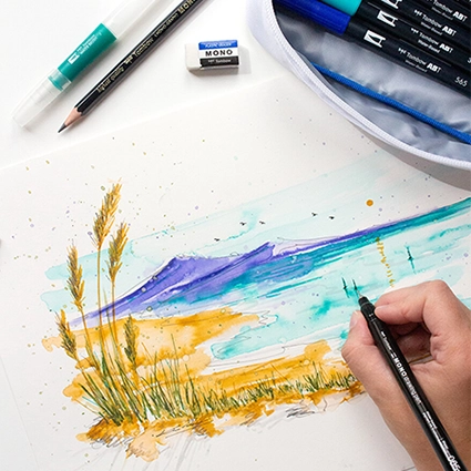Jemand malt mit Brush Pens von Tombow eine Landschaftsszene auf ein Blatt Papier. Ein Etui mit weiteren Stiften liegt daneben.