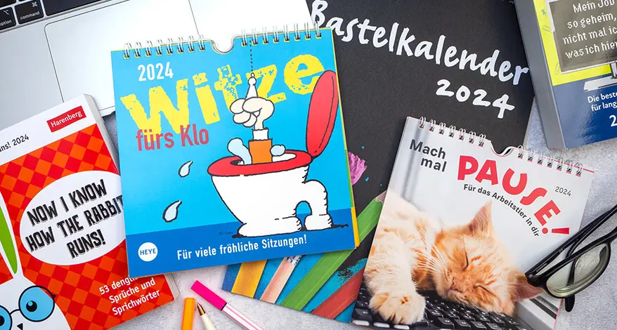 Viele verschiedene Kalender mit Witzen und lustigen Sprüchen vom Athesia Kalenderverlag liegen auf einem Schreibtisch verteilt. Foto: Johanna Rundel