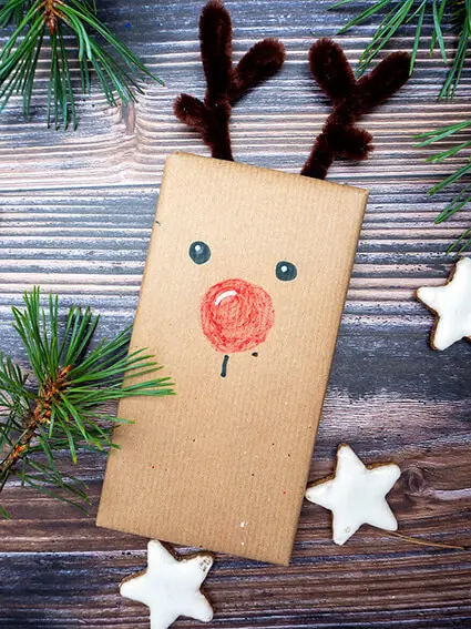 Auf einer rustikalen Holzplatte liegt eine weihnachtlich verpackte und mit Stiften von STAEDTLER als Rentier bemalte Schokoladentafel. Foto: Johanna Rundel