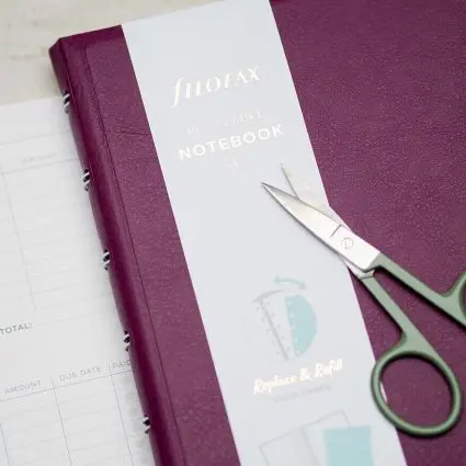 Auf einem weinroten Filofax-Notizbuch liegt eine kleine, salbeigrüne Schere. Foto: Johanna Rundel