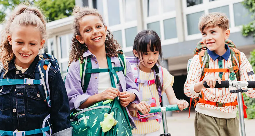 Eine Gruppe Schülerinnen und Schüler im Grundschulalter laufen mit ihren Schulranzen und Sporttaschen von ergobag nebeneinander über den Schulhof. 