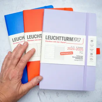 Jemand legt verschiedenfarbige Kalender, Planer und Notizbücher von LEUCHTTURM1917 auf einem grauen Schreibtisch aus. Foto: Johanna Rundel