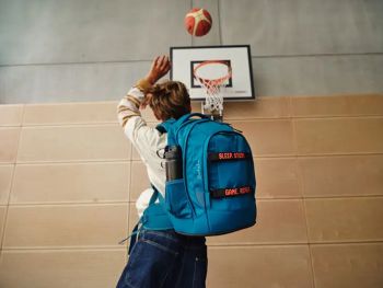 Ein Junge mit einem blauen Schulrucksack von satch auf dem Rücken wirft in der Sporthalle einen Ball in den Basketballkorb.