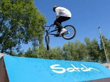 Ein Jugendlicher springt mit seinem Fahrrad über eine blaue Rampe, auf der in weißer Schrift „satch“ steht. 