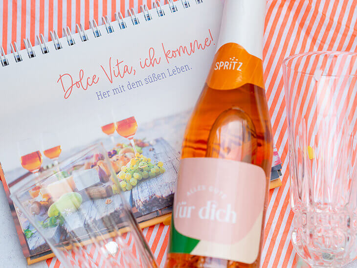 Ein Motivationsaufsteller mit Sprüchen zum Ruhestand und eine Geschenk-Flasche „Sprizz“ von Grafik Werkstatt liegen zwischen Sektgläsern auf einem orange-weiß gestreiften Geschenkpapier. Foto: Johanna Rundel