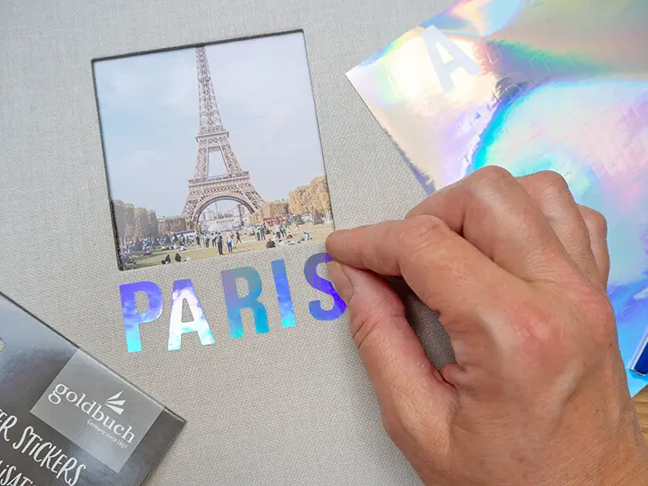 Jemand klebt das Wort „PARIS“ mit glitzernden Buchstabenaufklebern auf ein hellgraues Leinen-Fotoalbum von Goldbuch. Foto: Johanna Rundel