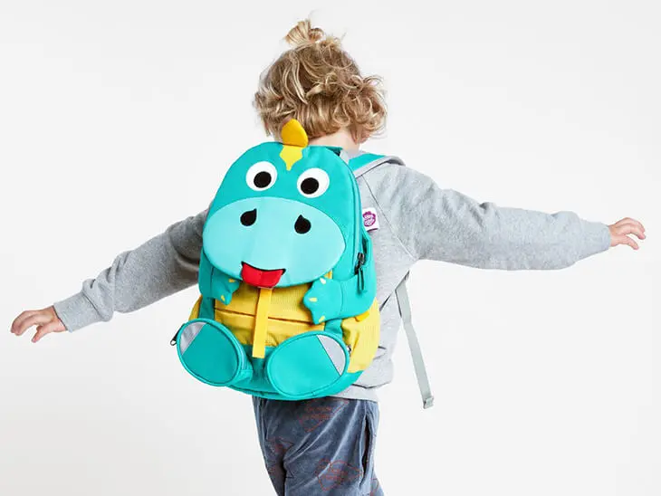 Ein Kindergartenkind tanzt mit einem Dino-Rucksack der Serie „großer Freund“ von Affenzahn vor einer weißen Wand.