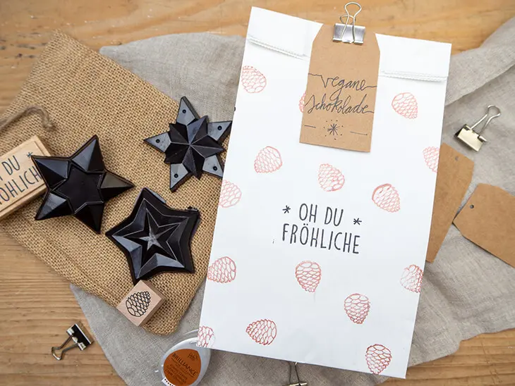 Eine mit weihnachtlichen Motiven bestempelte, weiße Papiertüte von HEYDA liegt mit selbstgemachter veganer Schokolade in Sternform auf einem weihnachtlich dekorierten Tisch. Foto: Johanna Rundel