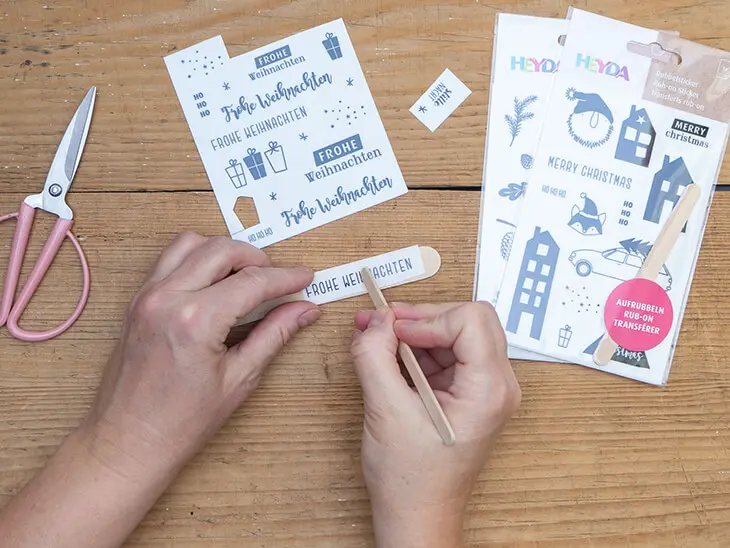 Jemand rubbelt einen Rubbel-Sticker von HEYDA auf einen Mundspatel aus Holz. Drumherum liegen weitere Bastelmaterialien. Foto: Johanna Rundel
