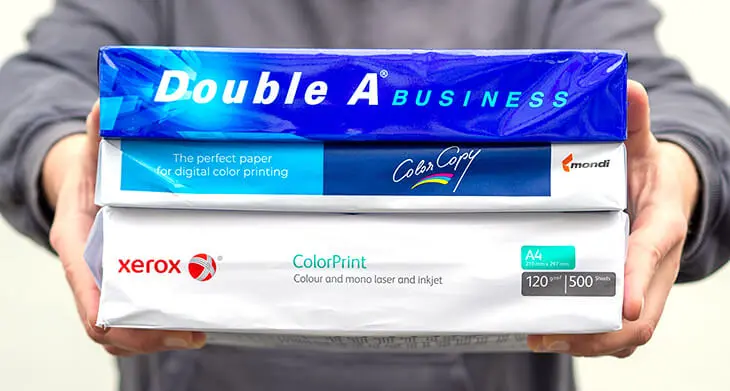 Eine Person in einem dunkelgrauen Pullover vor einer weißen Wand trägt drei verschiedene Packungen Kopierpapier vor der Brust. Foto: Johanna Rundel