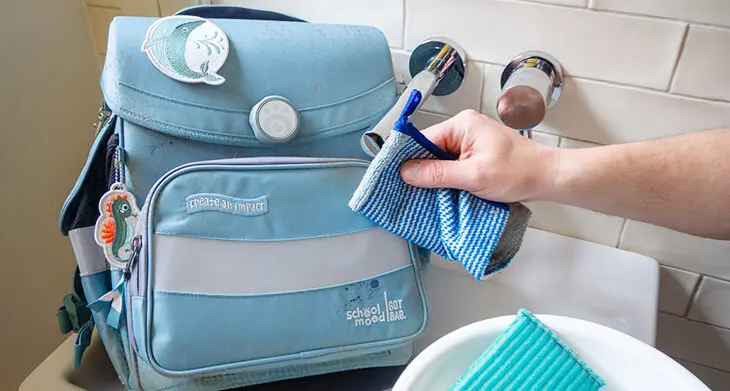 Jemand wischt mit einem blauen Schwamm die Außenseite eines blauen Schulranzens von School Mood sauber, der in einem Waschbecken steht. Foto: Johanna Rundel