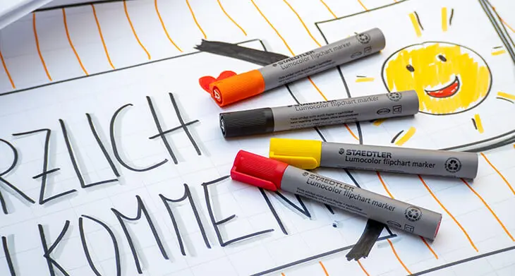 Verschiedenfarbige Flipchartmarker von STAEDTLER liegen auf einem bunt gestalteten Willkommens-Flipchart-Bogen mit einer gelben Sonne. Foto: Johanna Rundel