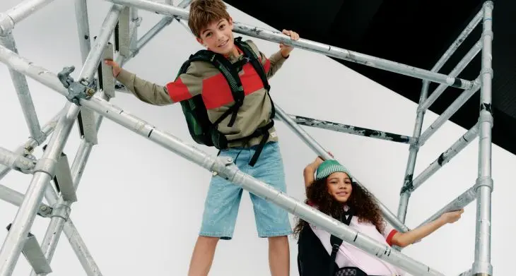 Zwei Jugendliche klettern auf einem Metallgerüst, beide tragen Schulrucksäcke von satch und lässige Freizeitkleidung