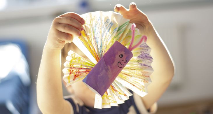 Ein Kind hält einen selbstgebastelten Schmetterling aus Klopapierrolle und bunt bemaltem Papier in die Kamera
