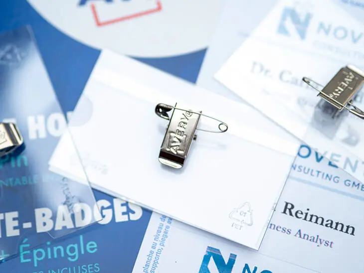 Ein Namensetikett mit Clip und Nadel von Avery Zweckform liegt zusammen mit weiteren Etikettenhüllen und Namensschildern auf einer hellen Unterlage. Foto: Johanna Rundel