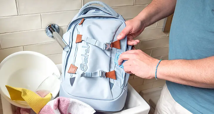Jemand steht an einem Waschbecken, in dem ein hellblauer satch Schulrucksack liegt und reinigt diesen mit einem Lappen. Daneben sind weitere Putzutensilien zu sehen. Foto: Johanna Rundel
