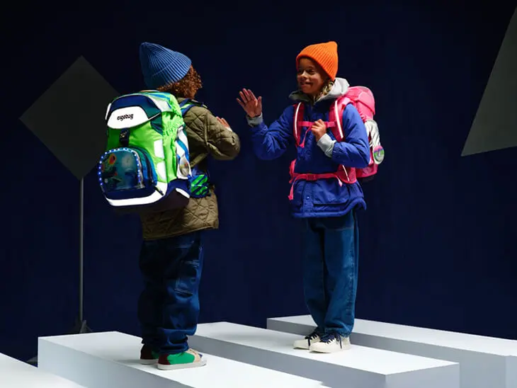 Zwei Kinder laufen mit reflektierenden Schulranzen von ergobag durch eine imaginäre Verkehrsszene.