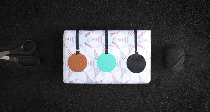 Auf einem schwarzen Untergrund liegt ein quadratisches Geschenk, welches in weißes Geschenkpapier mit Sternen eingepackt ist. Darauf sind mit braunem, türkisem und schwarzem Papier runde Kreise als Weihnachtskugeln an einem schwarzen Band aufgeklebt.