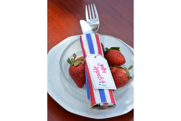 Auf einem Holztisch steht ein weißer Teller, auf diesem steht eine Glasschale gefüllt mit Erdbeeren. Besteck ist, eingewickelt in eine Serviette, darüber gelegt und mit einem Tafeletikett mit der Aufschrift "Guten Appetit" verzeirt.