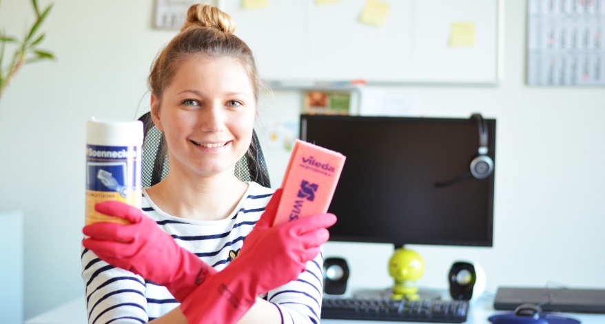 Eine Frau sitzt im Büro, trägt Gummihandschuhe und hält Reinigunsmittel in der Hand.