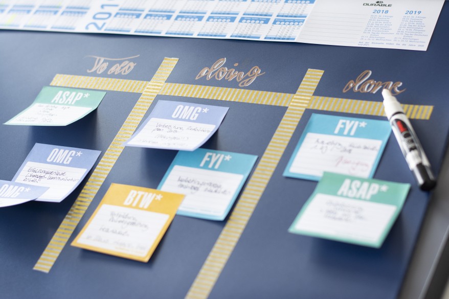 Auf eine blaue Schreibtischunterlage der Marke Durable ist ein Kanban Board gemalt. In den verschiedenen Rubriken to do, doing und done wurden verschiedene beschriftete Haftnotizen geklebt. Am Bildrand ist ein Kalender zu sehen. Auf der Schreibtischunterlage liegt ein geöffneter Chalkmarker in weiß.