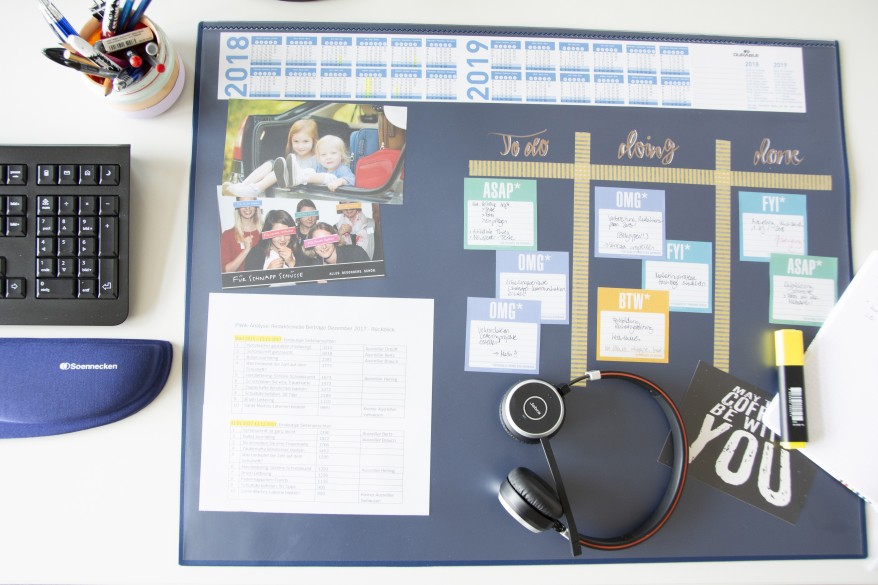 Auf einem weißen Schreibtisch sind eine schwarze Tastatur, eine Handauflage der Marke Soennecken sowie eine Schreibtischunterlage von Durable zu sehen. Auf der Unterlage liegen ein Headset und ein gelber Textmarker. Im oberen Bildrand befindet sich ein gefüllter Stifteköcher. In der Schreibtischunterlage sind verschiedene Zettel, Bilder sowie Kalenderübersichten für das Jahr 2018 & 2019. Außerdem wurde dort mit verschiedenen Haftnotizen ein Kanban-Board erstellt. 