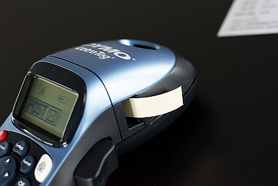 Dymo-Beschriftungsgerät mit herausragendem Etikettenband auf Tisch.