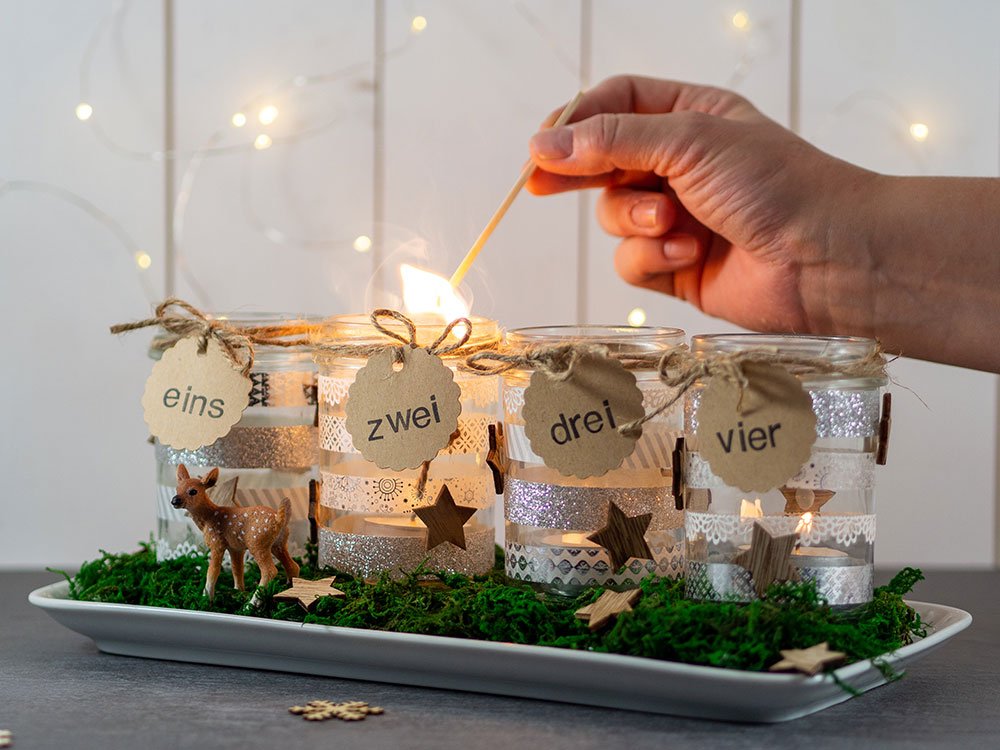 Ein selbstgebastelter Adventskranz steht auf einem grauen Tisch. Eine Hand hält ein brennendes Streichholz, das gerade eine Kerze anzündet. Der Hintergrund ist weiß und es brennt eine Lichterkette.